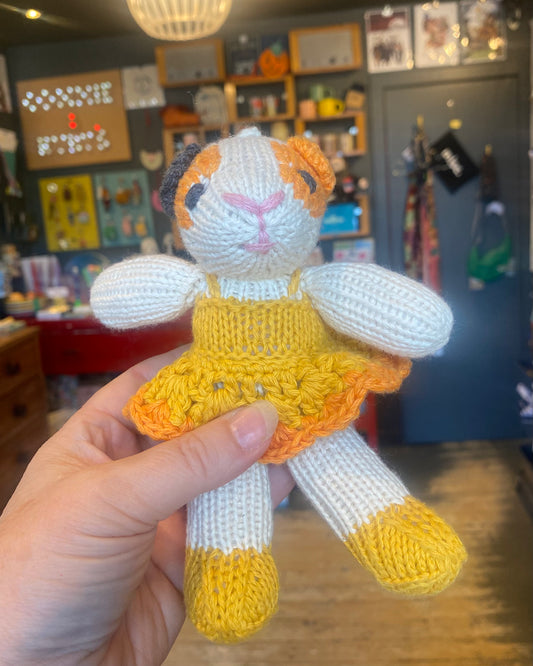 Handmade knitted Guinea pig toy with a yellow dress, held in front of a blurred indoor shop background.
