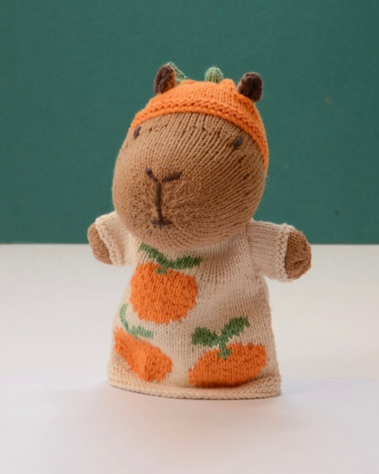 A hand-knit capybara puppet wearing an orange pumpkin hat and a cream sweater with orange pumpkins and green stems, standing against a teal background.