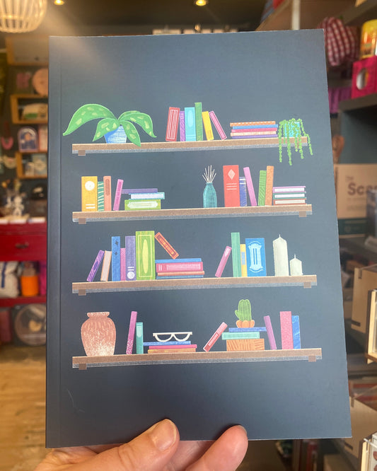 A hand holds a dark slate blue notebook featuring an illustration of colourful books on wooden shelves, with a potted plant, vase, and cactus. The background shows a blurred bookstore interior with shelves and lighting.
