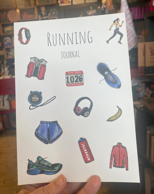 A hand holds a white running journal cover featuring hand-drawn illustrations of running gear and a race bib, with the title 'RUNNING JOURNAL' printed at the top, set against a blurred shop background.