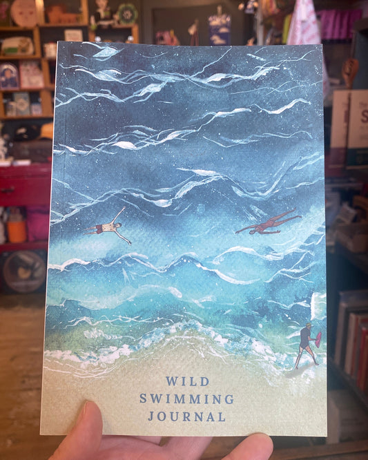 A hand holds a journal titled 'WILD SWIMMING JOURNAL' featuring an artistic illustration of swimmers in a blue ocean. The background shows a cozy, cluttered bookshop interior.