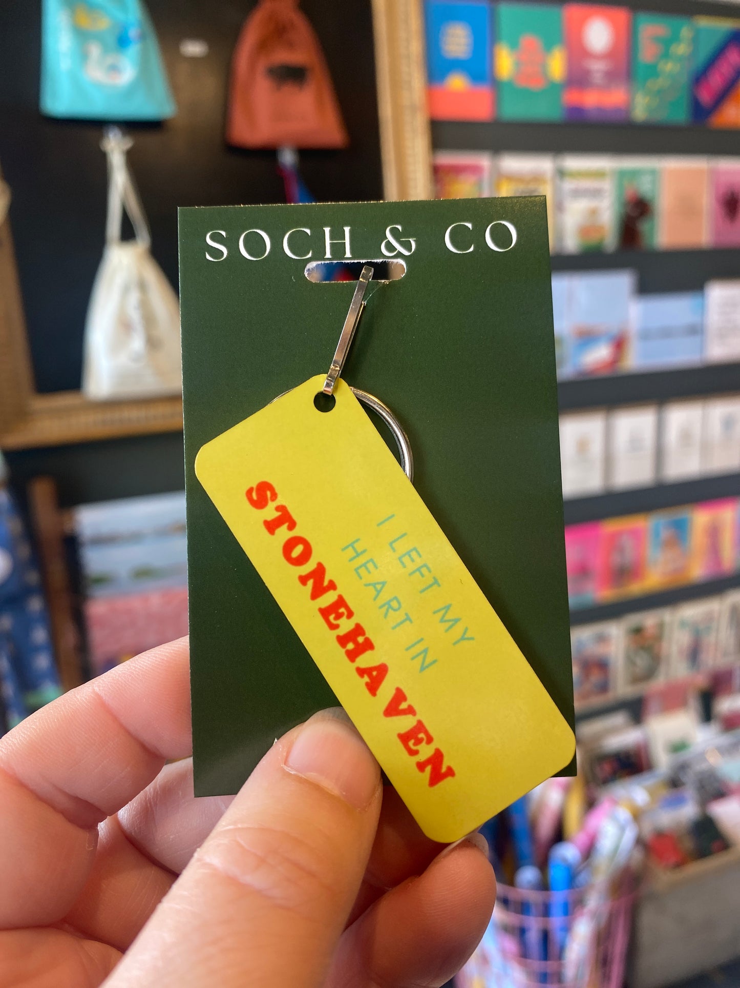 A hand holding a yellow rectangular keyring attached to a dark green card. The tag reads 'I LEFT MY HEART IN STONEHAVEN' in red and blue text.