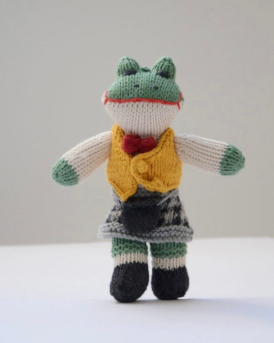A hand-knitted frog doll wearing a yellow vest, red bow tie, and a dark patterned skirt, standing against a plain light grey background.