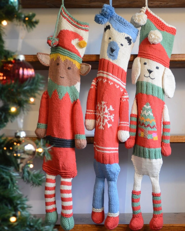 Three knitted Christmas stockings with elf, bear, and dog designs hanging on a fireplace.