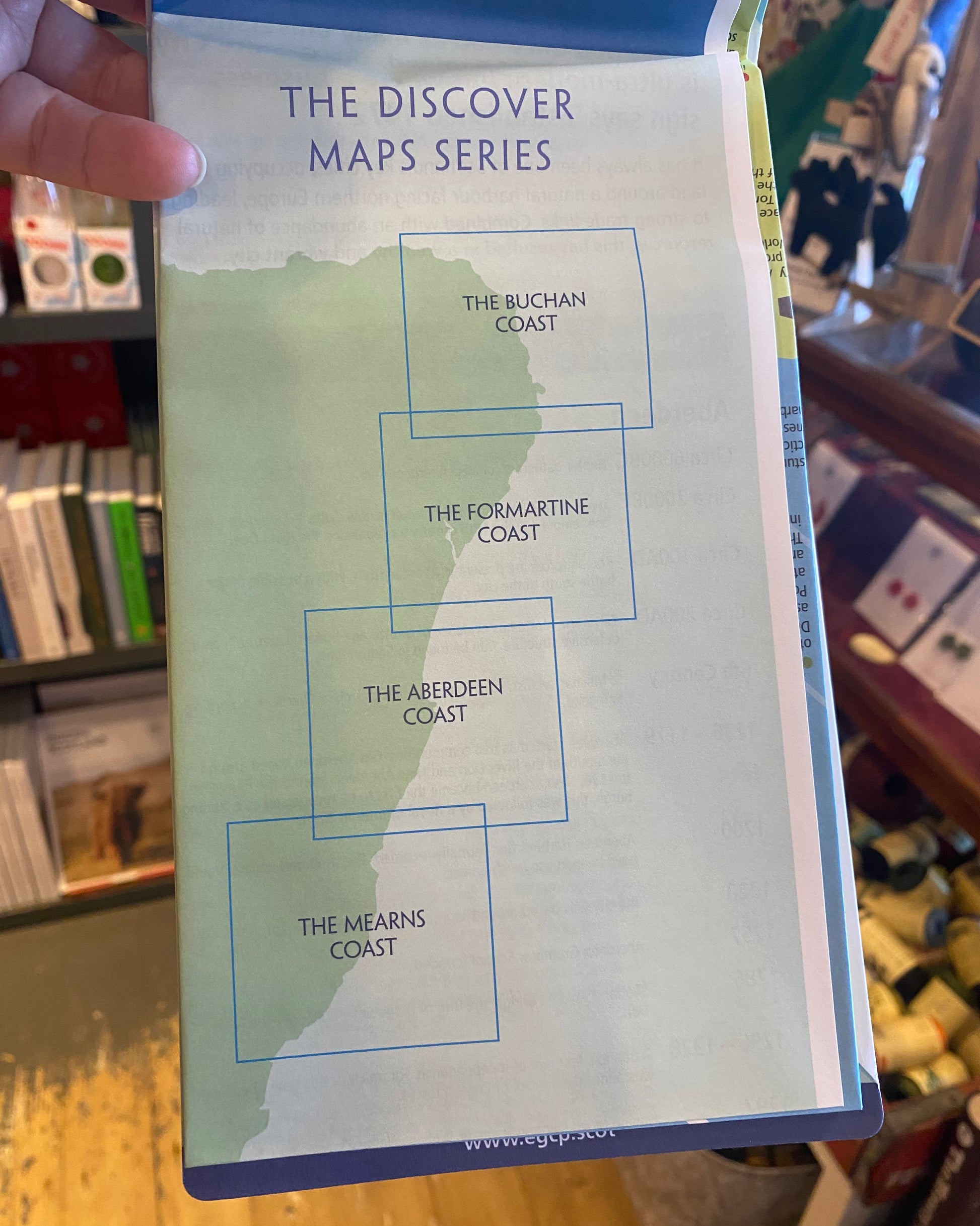 Map titled 'The Discover Maps Series' open to shop different areas covered, held by a hand with a store interior in the background.