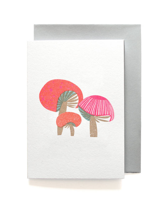 A white greeting card featuring a minimalist illustration of two pink and coral mushrooms, shown with a silver-gray envelope behind it on a clean white background.