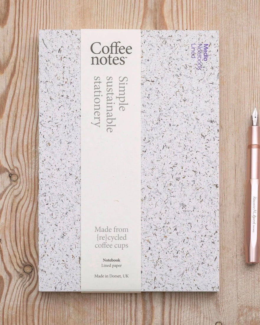 A notebook with a white and grey speckled seaweed cover and a central cream-coloured label reading 'Coffee notes' and 'Simple sustainable stationery,' placed on a light wood surface next to a rose gold fountain pen.