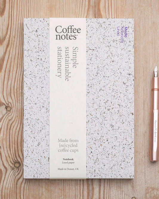 A notebook with a white and grey speckled seaweed cover and a central cream-coloured label reading 'Coffee notes' and 'Simple sustainable stationery,' placed on a light wood surface next to a rose gold fountain pen.