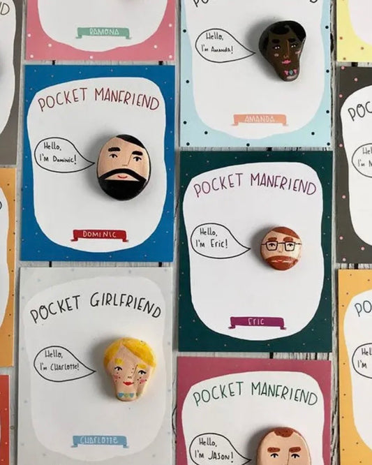 A close-up of several small painted stone faces mounted on colourful backing cards. The cards read phrases like "POCKET MANRIEND," "POCKET GIRLFRIEND," and "Hello, I'm [name]!" with illustrated speech bubbles.