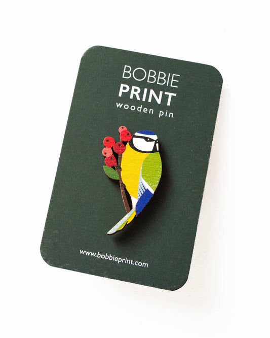 A wooden pin shaped like a blue tit bird perched on a branch with red berries, attached to a dark green backing card printed with 'BOBBIE PRINT wooden pin' and the website, photographed on a white background.