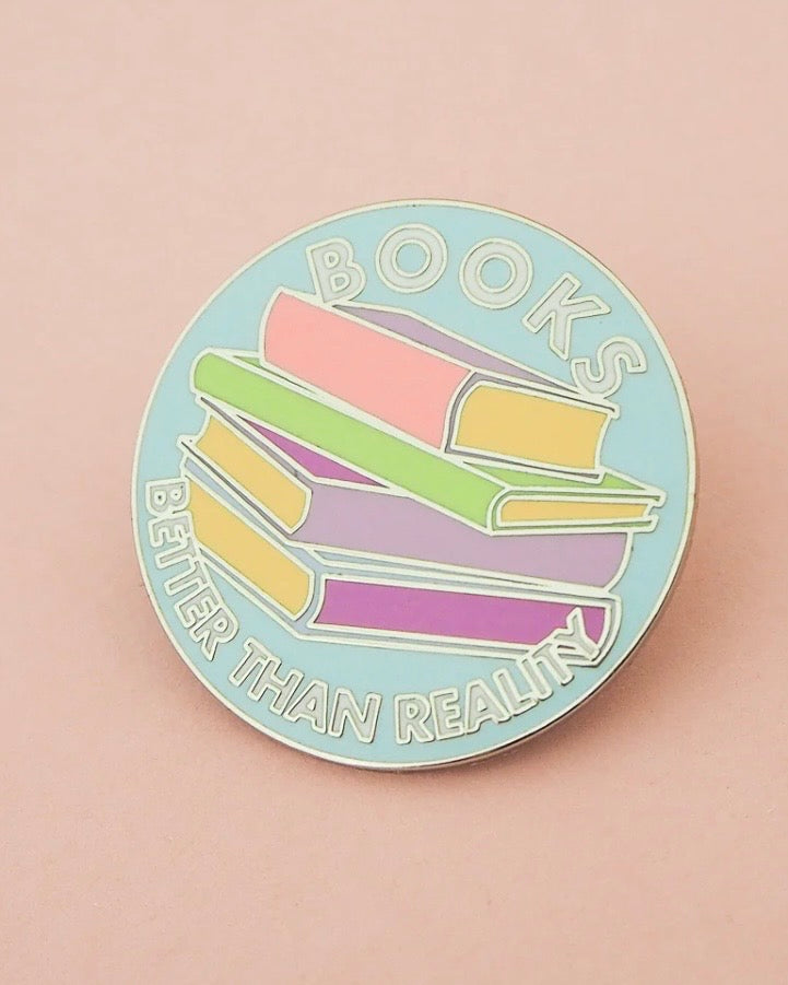A round enamel pin featuring a stack of pastel-colored books with the text 'BOOKS BETTER THAN REALITY' on a light pink background.