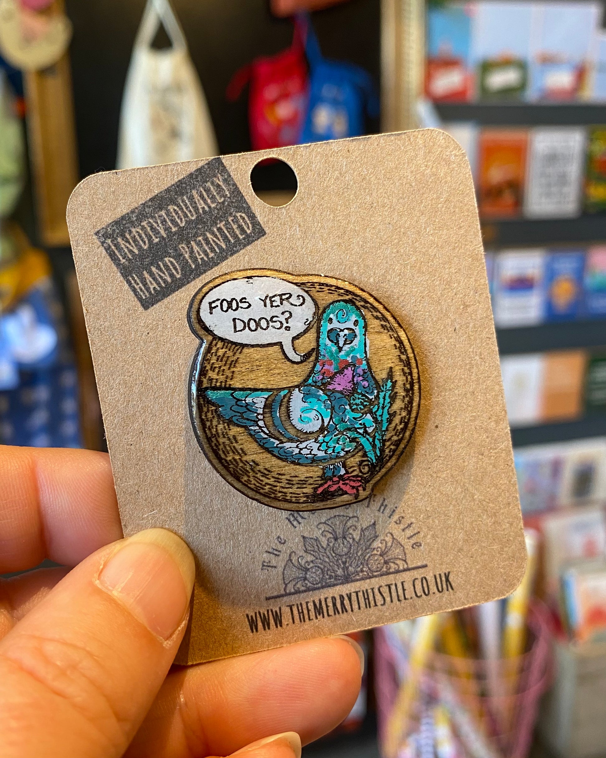 Pin with a pigeon character and speech bubble reading ‘Foos yer Doos?’ on a brown card, held in front of a store shelf.