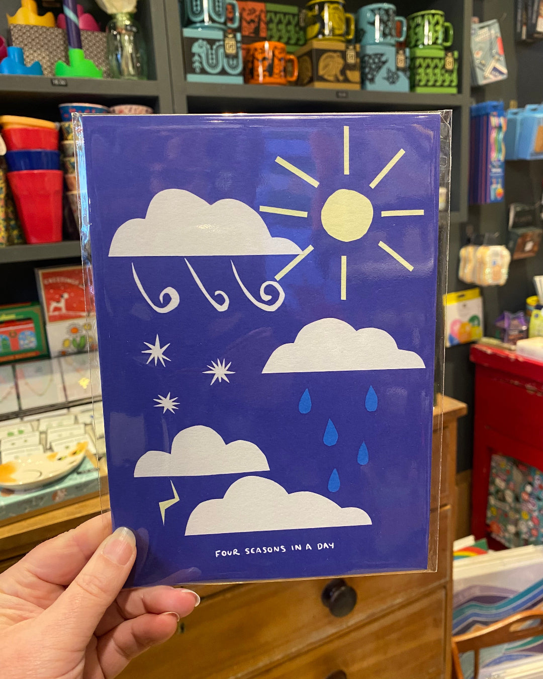 Blue A5 print with weather illustrations held in a hand, with a store shelf in the background.