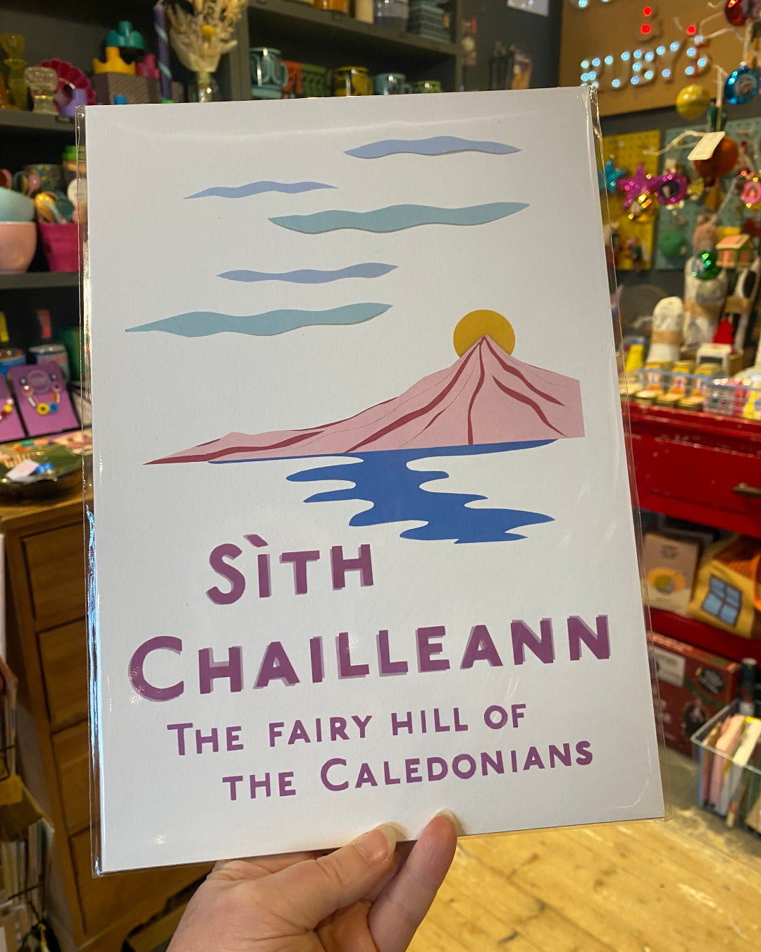 Print with illustration of a mountain, Schiehallion, and Gaelic text held in front of a store shelf.