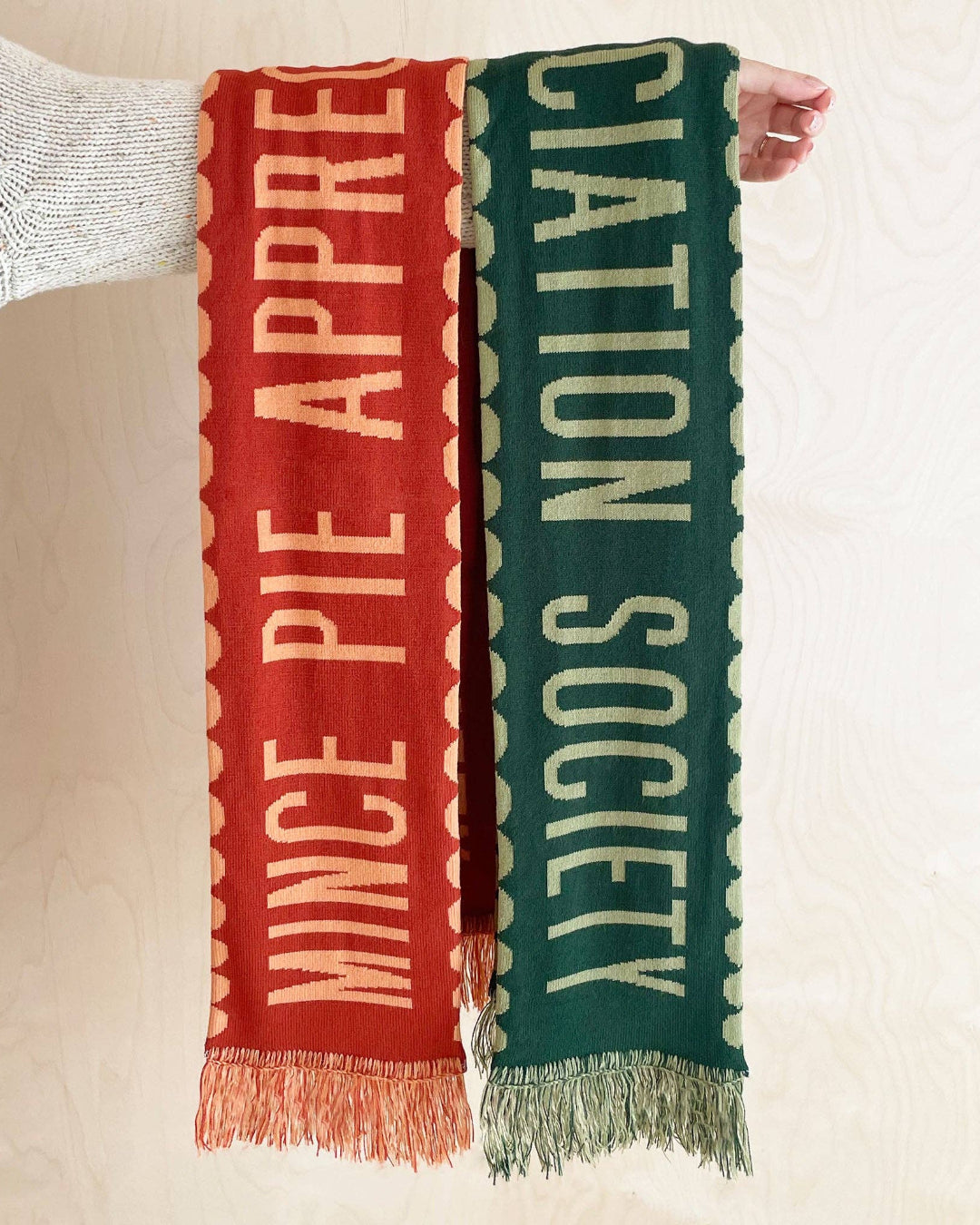 Two scarves with text, one red and one green, held against a neutral background.