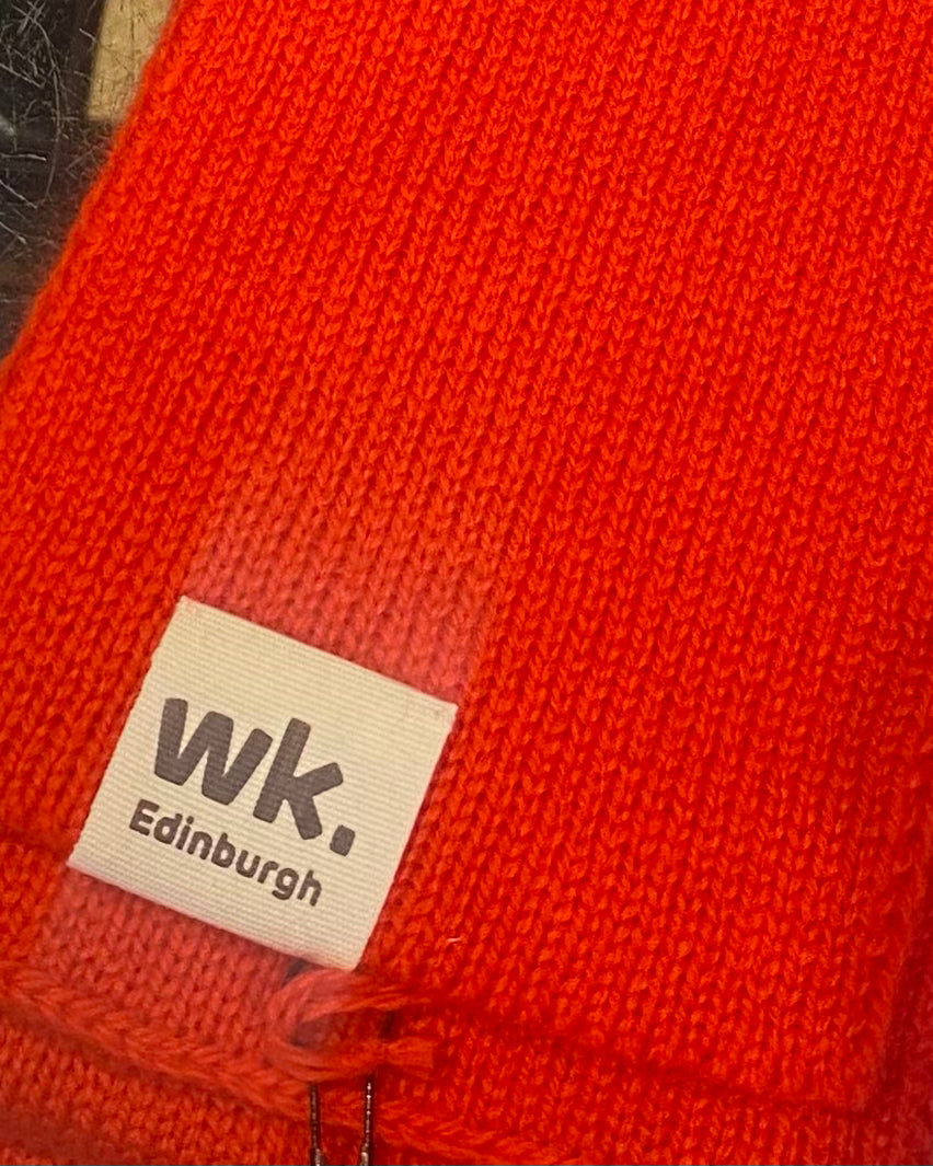 Close-up of a red merino wool scarf with a beige label featuring 'wk. Edinburgh'.
