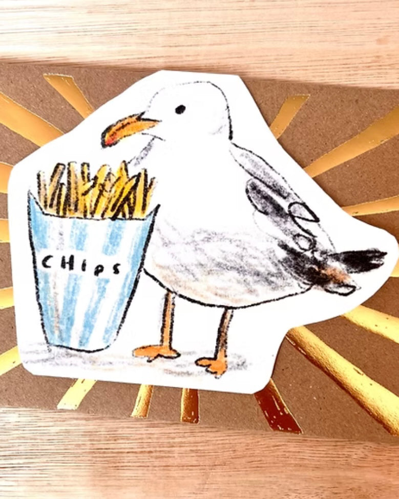 Shaped card with an illustration of a seagull holding a bag of chips on a foiled envelope, on a wooden background.