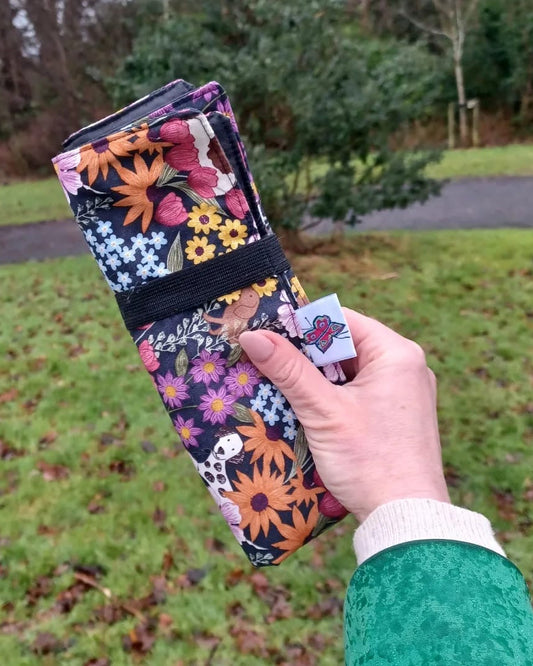 A hand holding a rolled-up sitting mat outdoors on a grassy path. The mat has a dense multicolour floral m pattern on a black background, secured with a black elastic band.