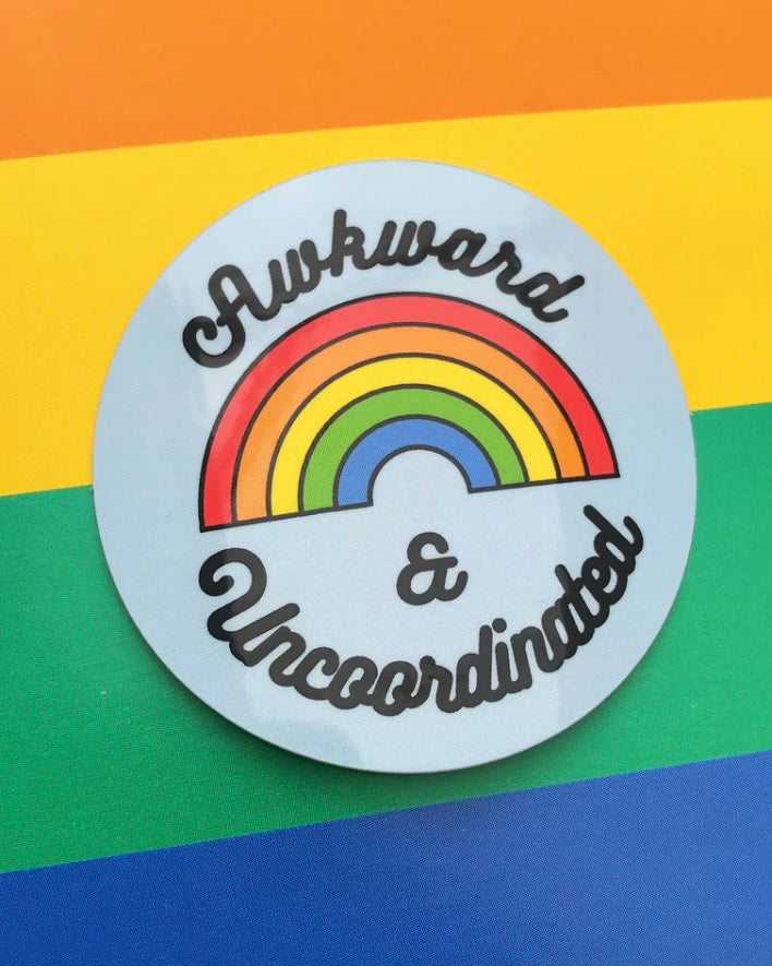A round sticker with a rainbow graphic and the text 'Awkward & Uncoordinated' in black script, shown against a rainbow-striped background.