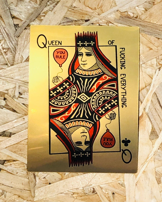 A square gold-toned sticker featuring a stylised queen playing card illustration with black, red, and orange details, placed on a bed of light wood shingles. The card reads 'QUEEN OF FUCKING EVERYTHING' and includes small red balloons with text.