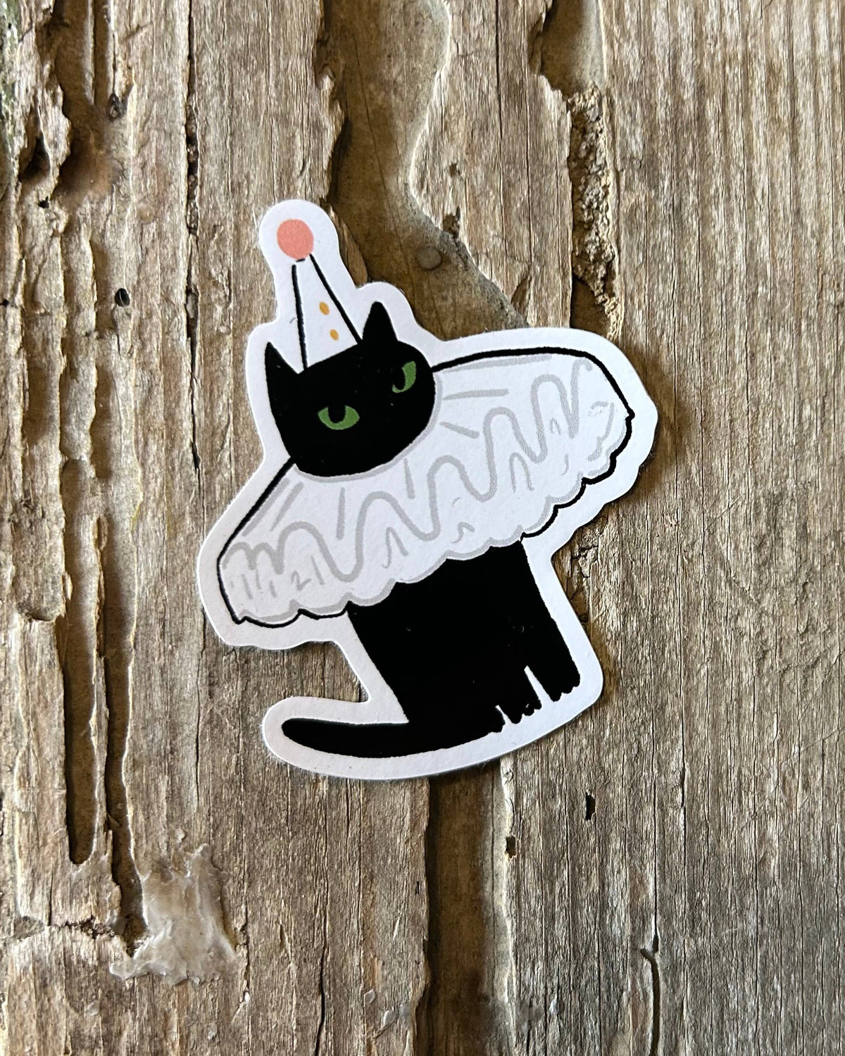 Sticker of a black cat in a party hat with a neck ruff, on a wooden surface
