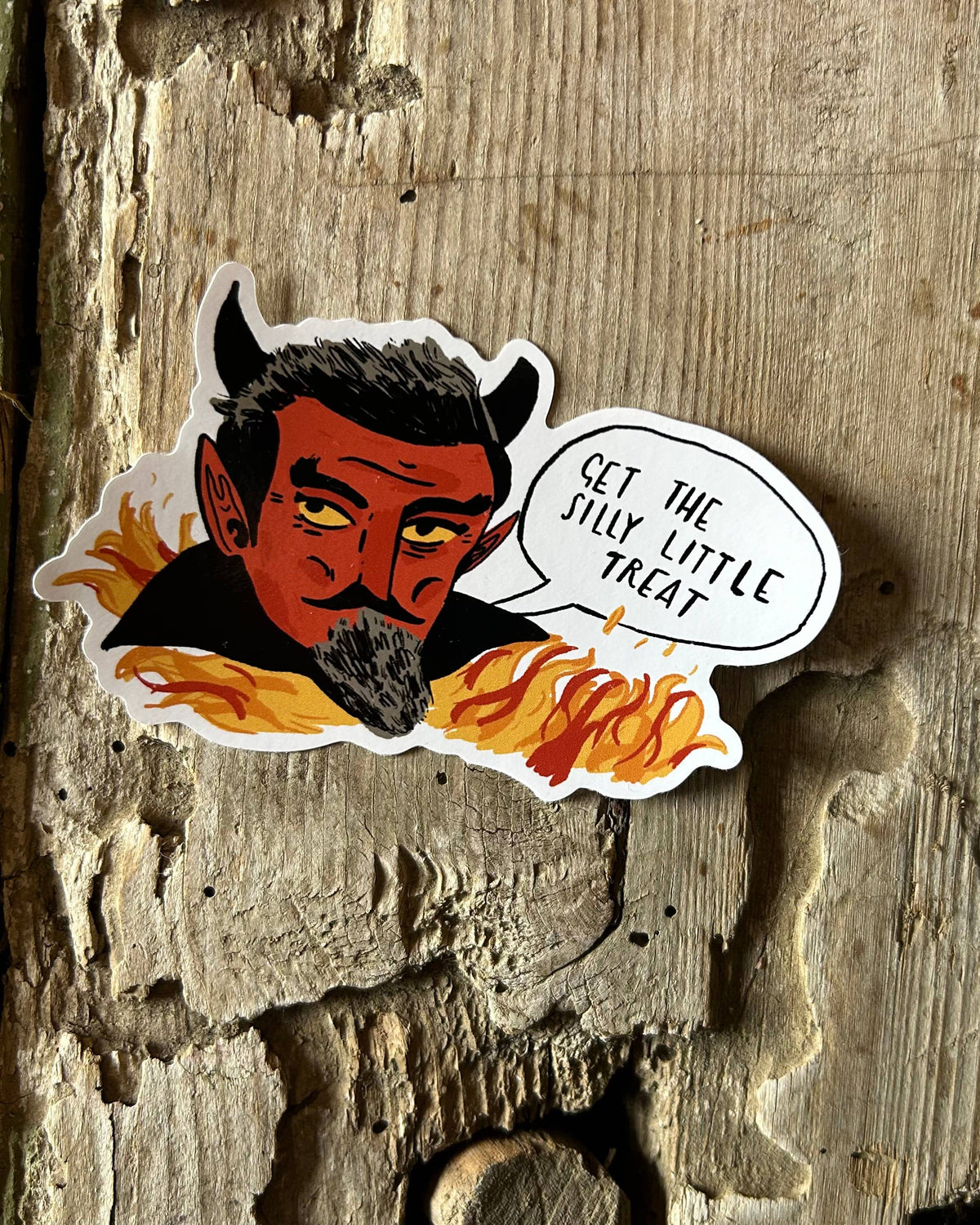 A die-cut vinyl sticker featuring a cartoon devil character with horns, yellow eyes, and flames, with a speech bubble reading 'GET THE ILLY LITTLE TREAT', shown on a rustic weathered wood background.