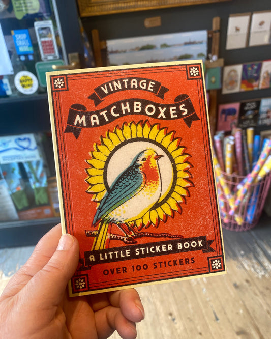 A hand holding a vintage-style matchbox sticker book cover labelled 'Vintage Matchboxes' with a bird illustration and text reading 'A Little Sticker Book' and 'Over 100 Stickers.' The background shows a blurred shop interior with shelves and colorful items.