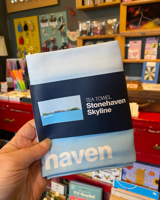 Hand holding a folded Teatowel with a belly band showing that it is of Stonehaven’s skyline, in a store setting.