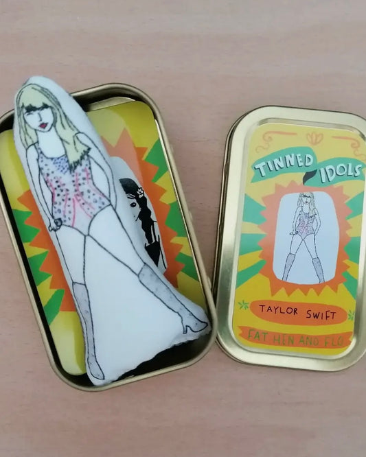 Open metallic tin with illustration of Taylor Swift  and text 'Tinned Idols Taylor Swift', with sewn mini Taylor inside, on a wooden surface.