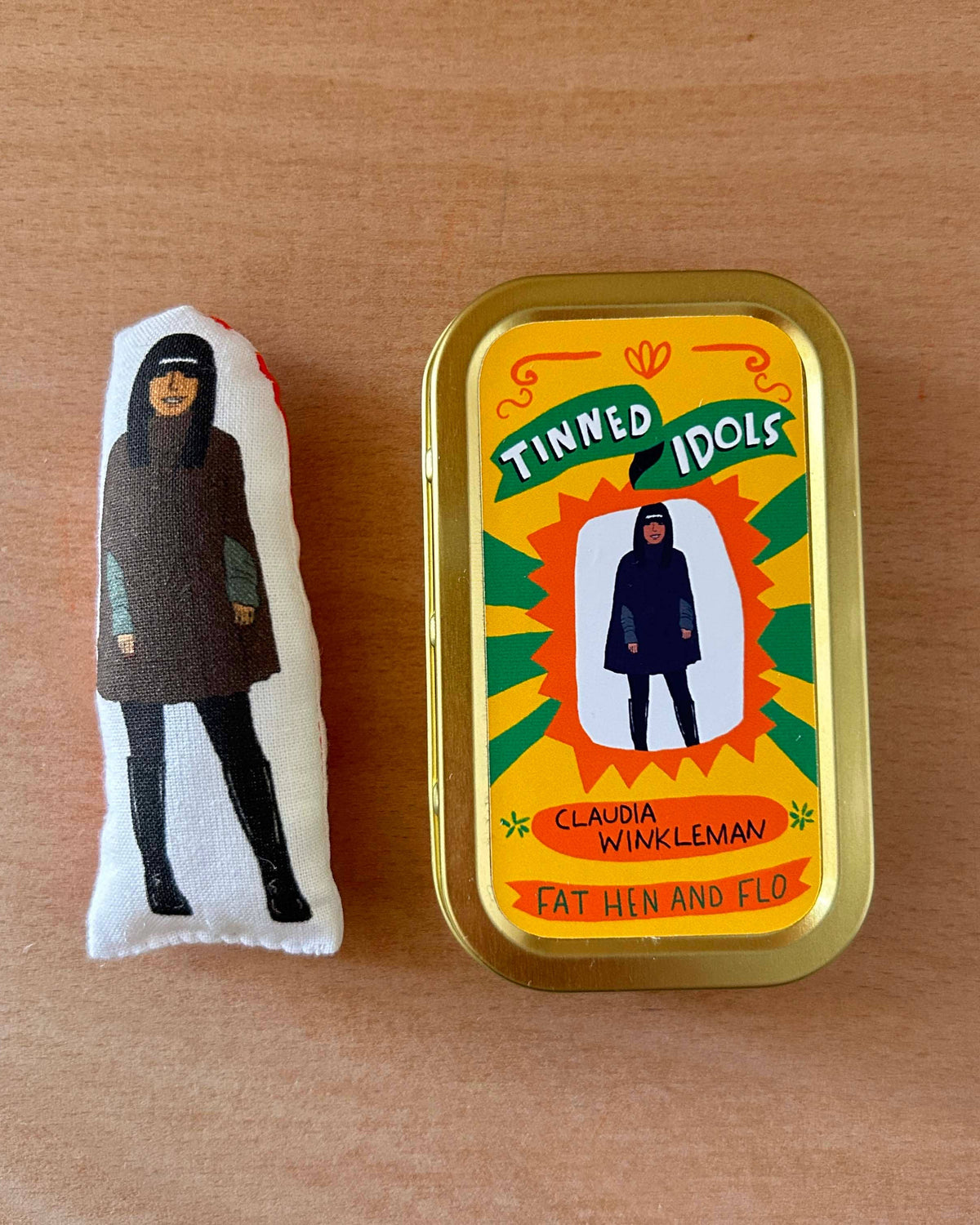 Small plush toy and tin with 'Tinned Idols' branding on a wooden surface