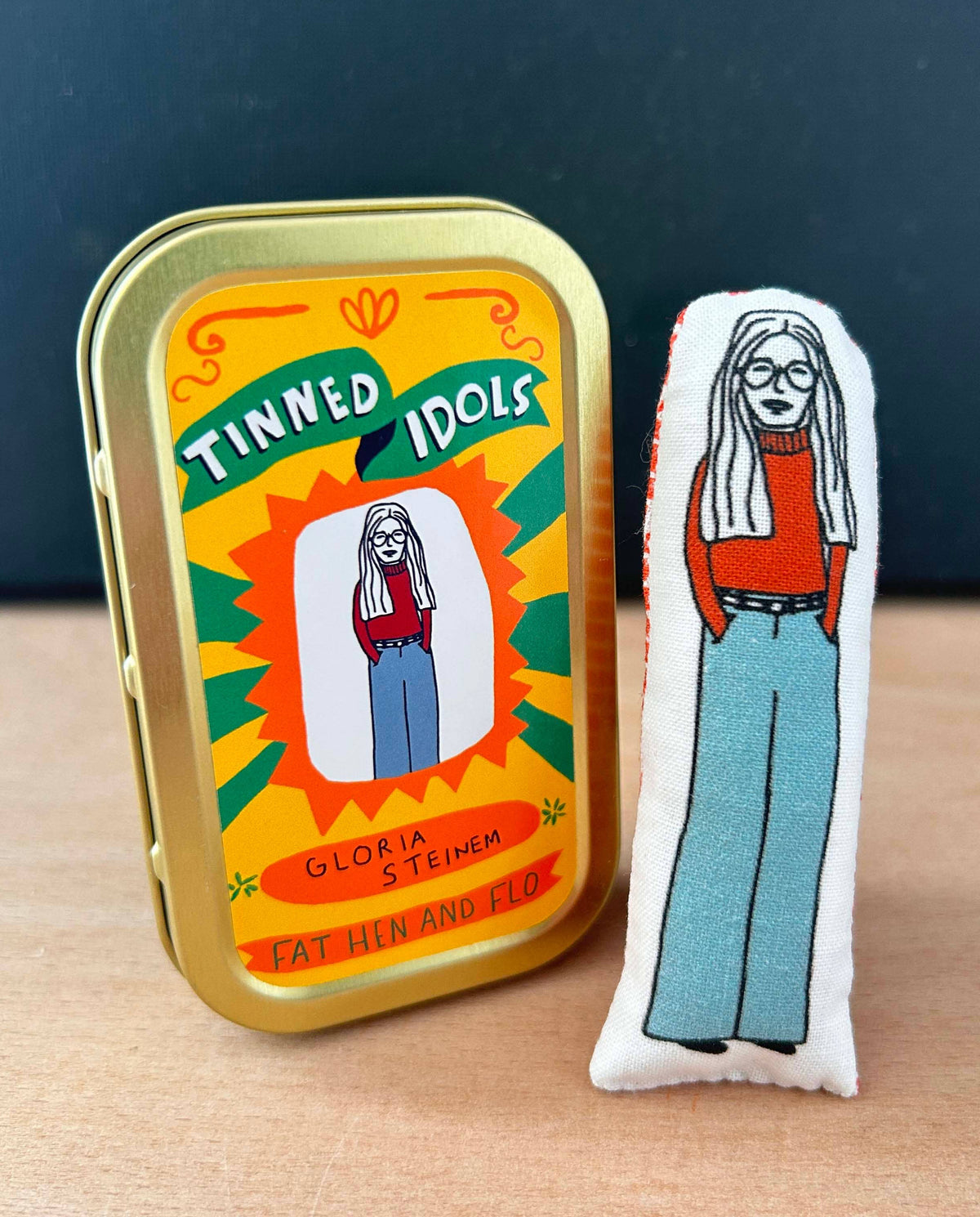 A gold metal tin with a retro 'Tinned Idols' label stands next to a white fabric pin featuring an illustrated woman in a red sweater and light blue pants, set on a wooden surface against a dark blue background.