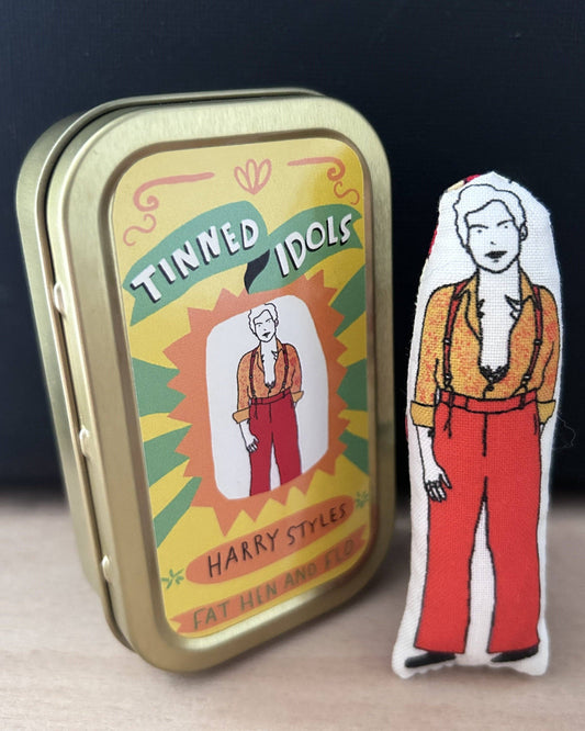 Metal tin labeled 'Tinned Idols' with a plush toy of a person wearing red pants and an orange jacket on a dark background.