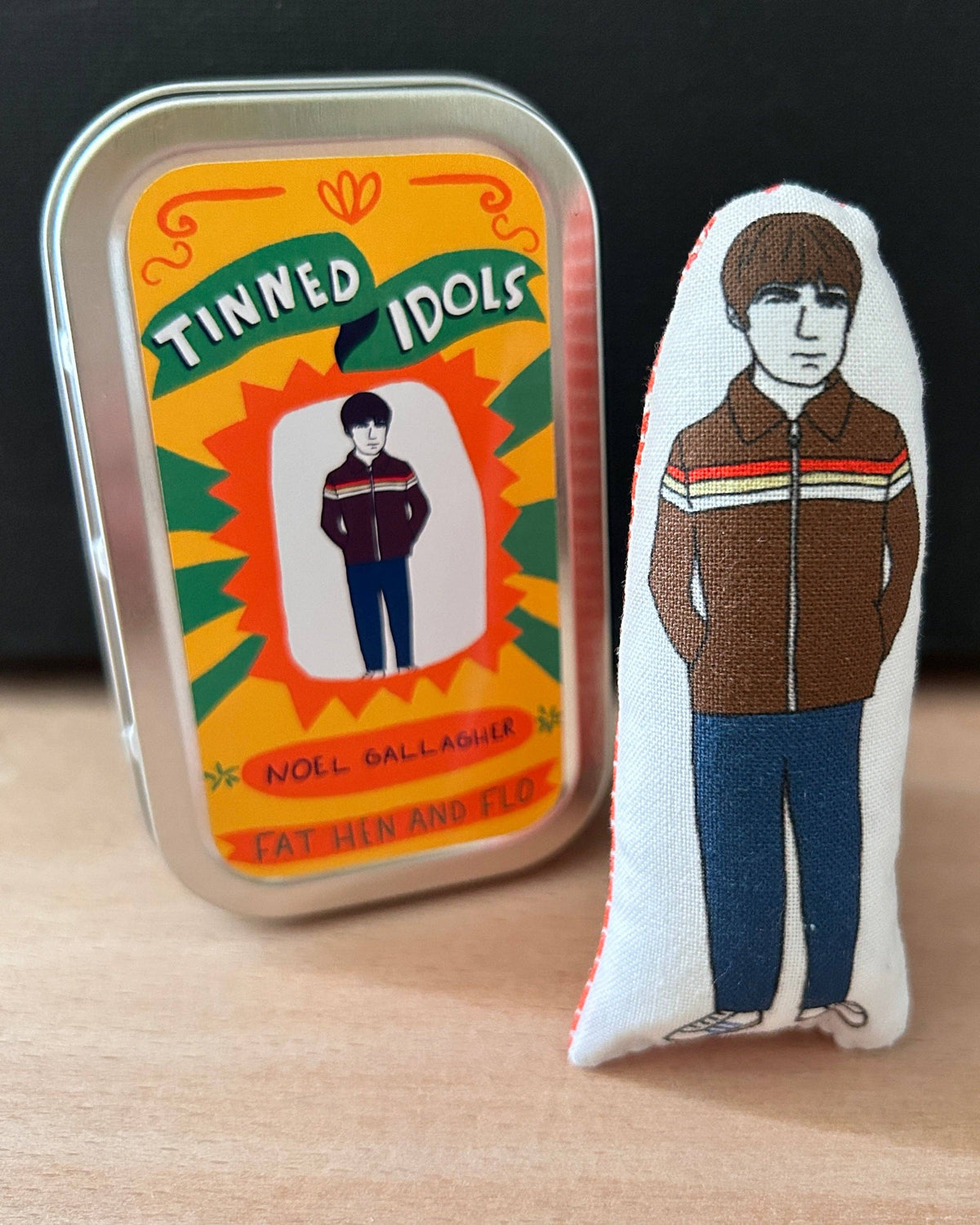 A small embroidered fabric figure of Noel Gallagher standing next to its original tin packaging on a light wood surface with a dark background.