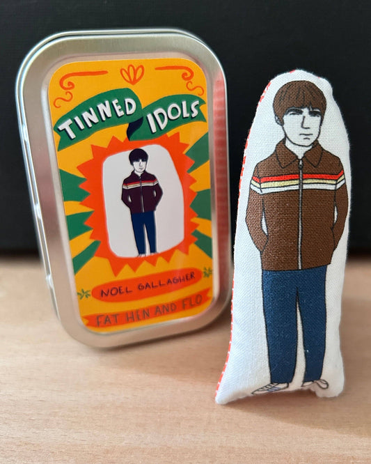 A small embroidered fabric figure of Noel Gallagher standing next to its original tin packaging on a light wood surface with a dark background.