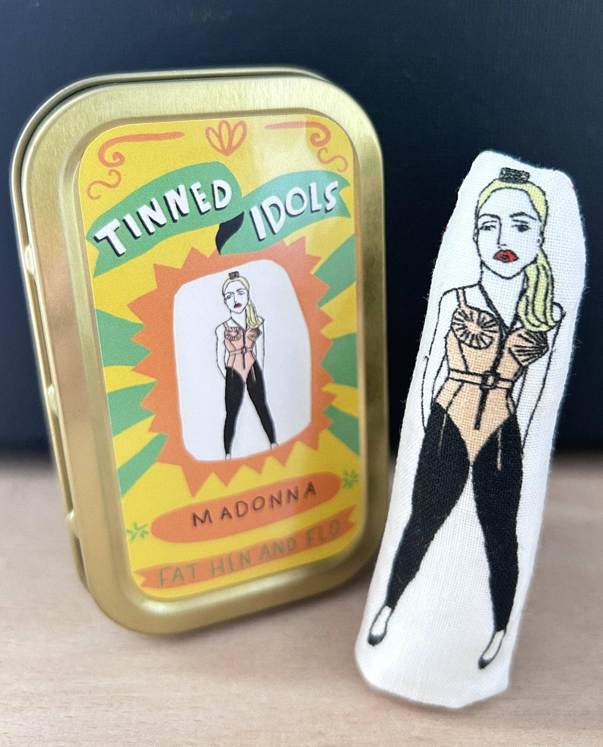 A small gold metal tin labeled 'Tinned Idols' with 'Madonna' and 'Fat Hen and Flo' printed on the front, shown next to a white fabric patch featuring an illustrated version of Madonna in a black and beige outfit, set on a light wood surface against a dark background.