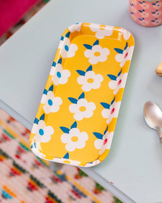 A yellow rectangular tray with a white floral pattern sits on a light blue table, styled with a pink mug, crackers, and a spoon in a cozy indoor setting.