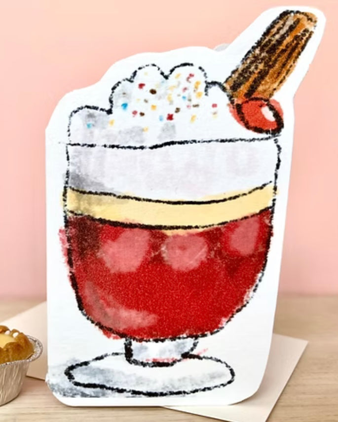 Shaped card with a trifle illustration, standing on an envelope, on a wooden table.