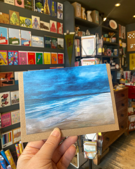Small square greetings card featuring a  painting of a blue ocean scene - on Barra - in a store with books and cards.