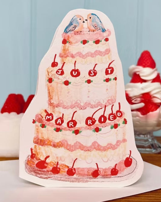 Decorative cake-shaped card with 'Just Married' text and cherries, on an envelope, with a light blue background.
